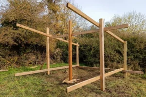 An upright wooden shelter frame in a field by a hedge