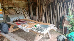Inside a barn with fence posts leaning on the back wall and a table with scything tool parts laid out