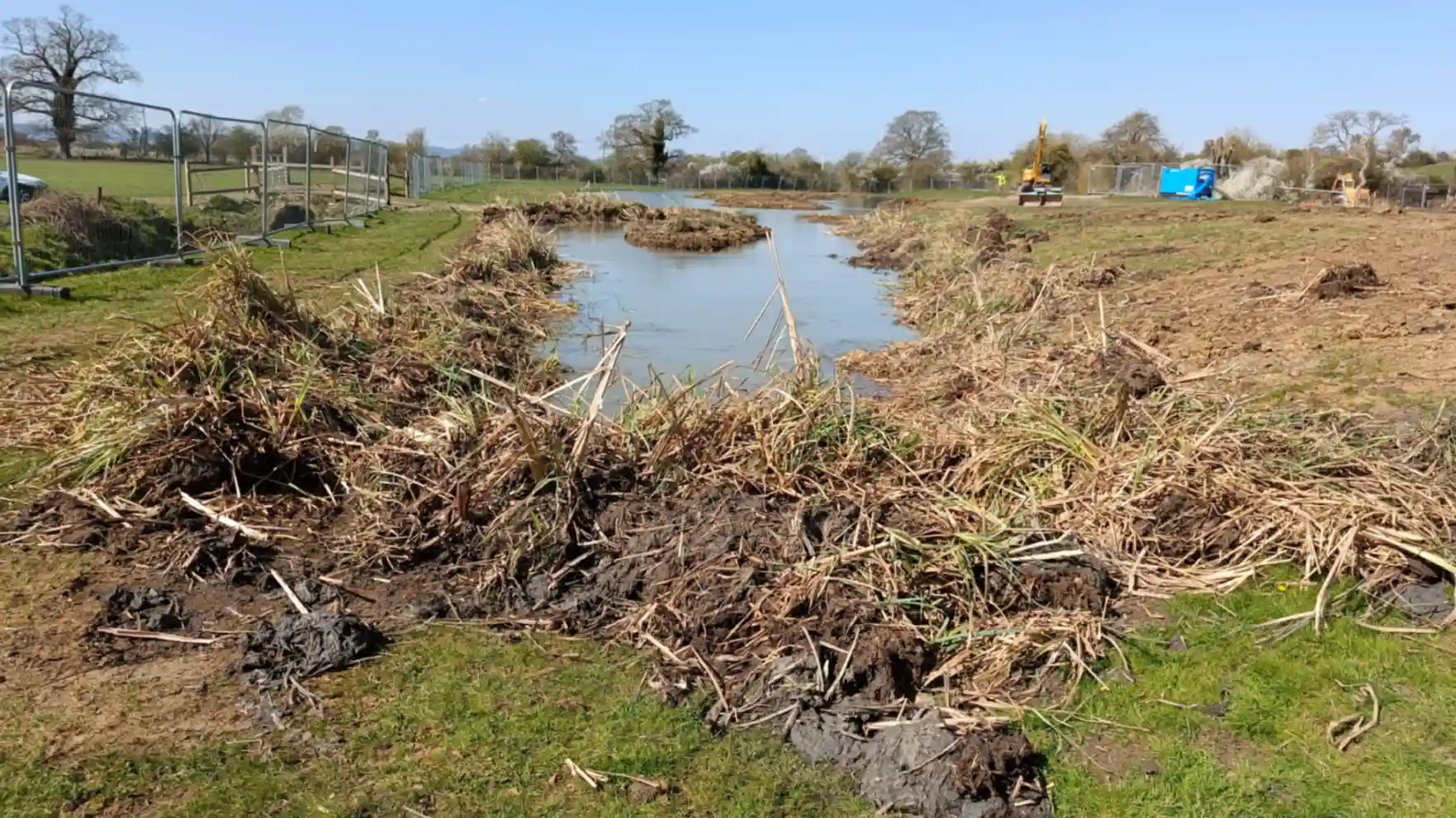 A large pond in a field with vegetation in the foreground and construction equipment in the background