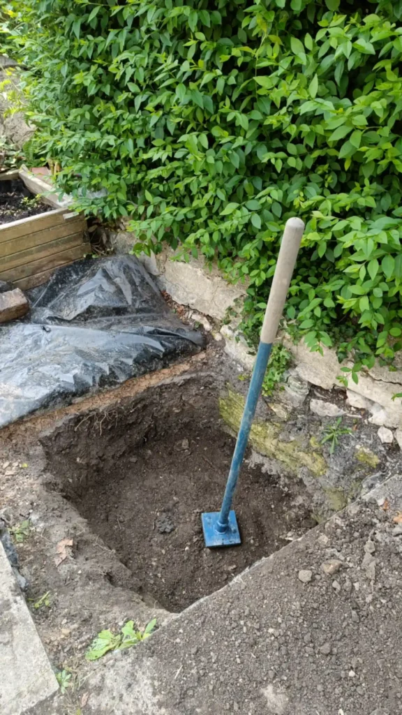 A pit half full with soil next to a hedge with a tamper stood up in it